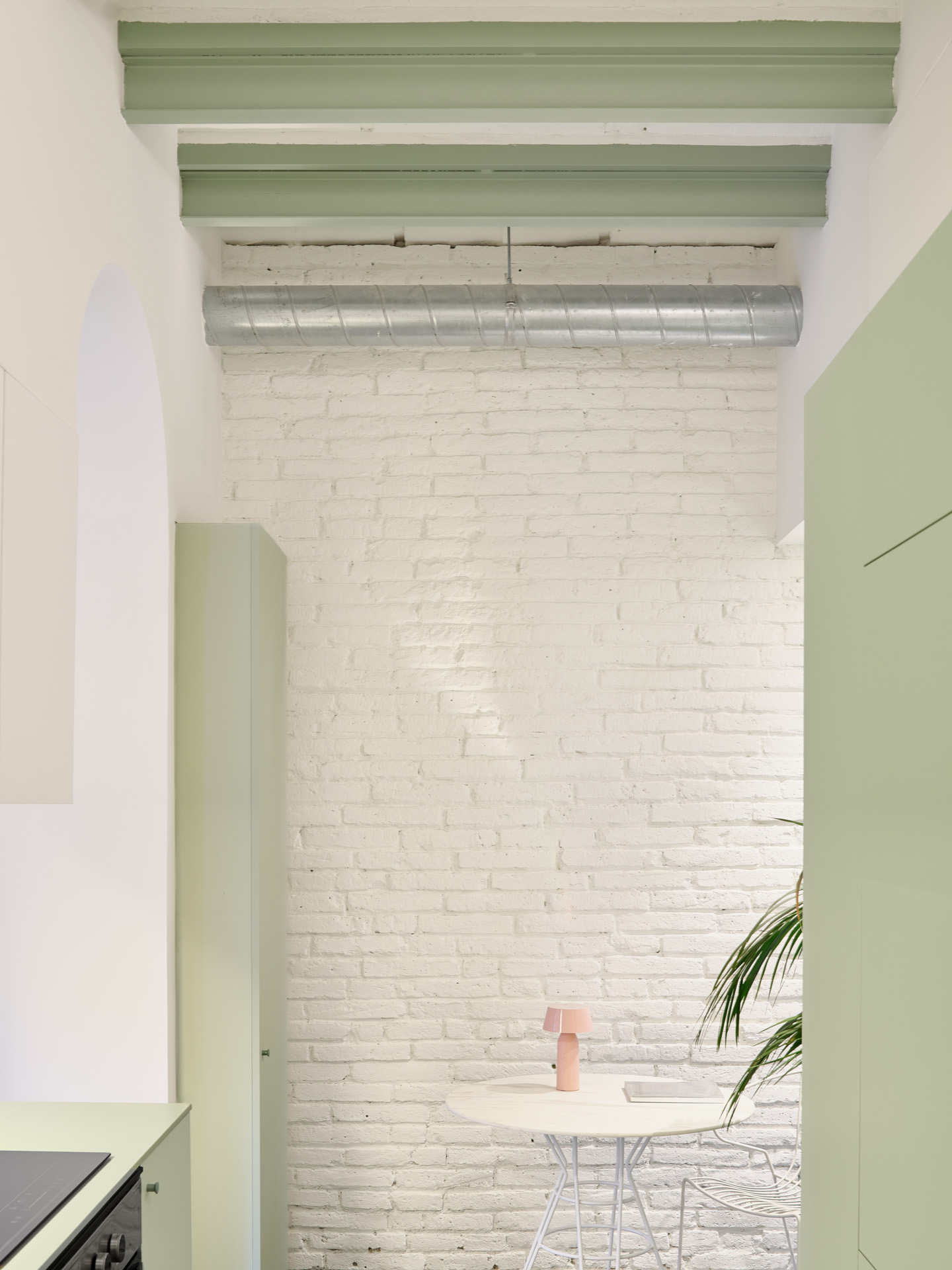 Dining nook in CRÜ’s “La Dolors” renovation, with a round white table, wire chairs, a whitewashed brick wall, and exposed green structural beams with a ventilation duct.